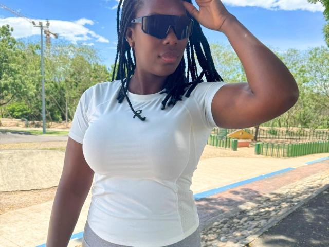 SaraMarks with dark skin, black braided hair, white tight shirt showing her big tits, grey pants, black sunglasses, and a necklace, standing outside on a sunny day.