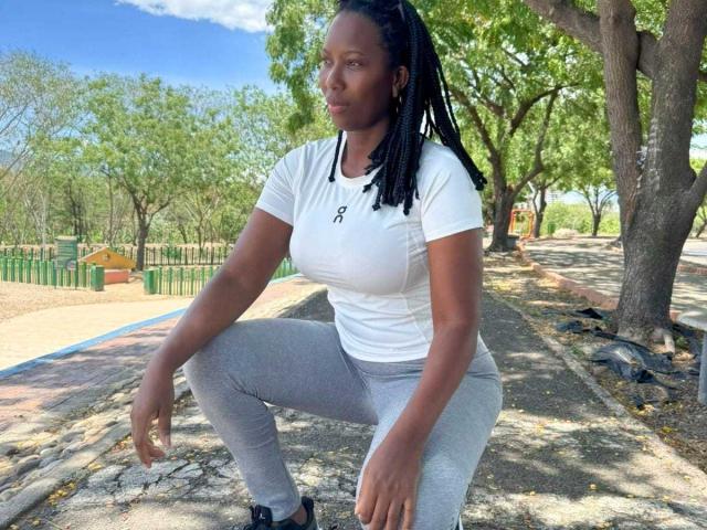 SaraMarks, squatting, black woman, medium dark skin, white tee, grey pants, black sneakers, long braids, small black tattoo on chest, green park, blue sky, trees, daytime.