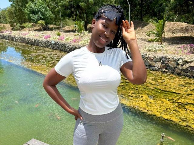 SaraMarks with dark skin, braided hair, white t-shirt, gray pants, standing by a green, rocky stream, smiling, hand on head, showing her boobs.