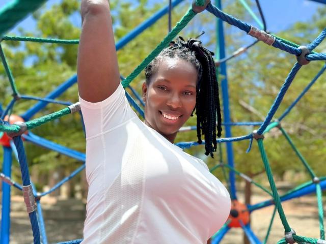 SaraMarks with dark skin, black braided hair, white shirt, smiling, arm raised, showing her tits, standing in a blue and green metal playground structure on a sunny day.