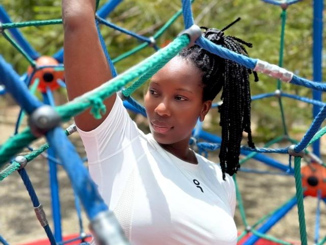 SaraMarks with dark skin, braided hair, white shirt, arm raised, smiling, blue and green rope climbing structure, her tits not visible, outdoor, daytime, greenery in background.
