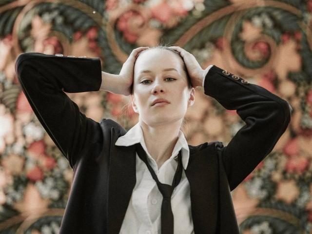 LynneLapora with hands in her pale, short hair, wearing a black blazer over a white shirt with a loose black tie, looking serious. Her tits are small.