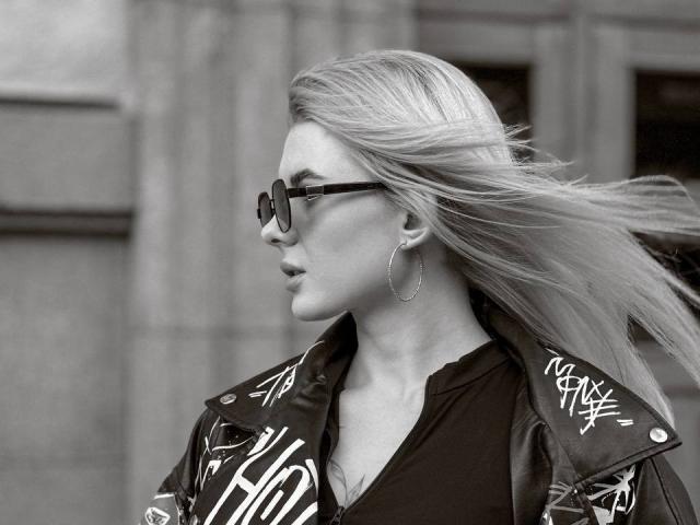 CynthiaMilkowski with long blonde hair, glasses, hoop earrings, and a graffiti-covered jacket, blowing in the wind, looking to the side. Black and white.