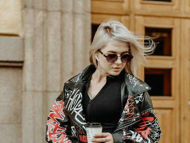 CynthiaMilkowski with blonde hair, wearing a black top, red and white graffiti leather jacket, and dark sunglasses, holding a coffee cup, wind blowing through her hair.
