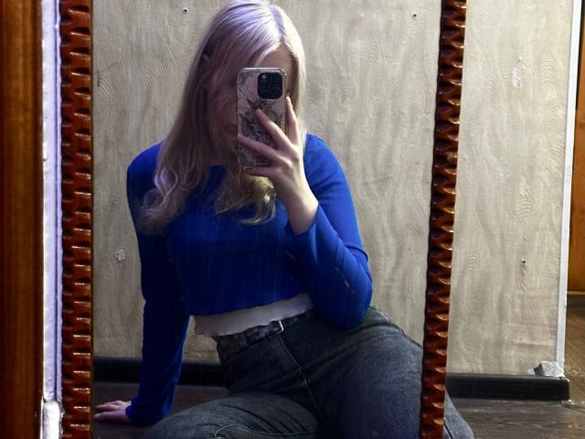 TiffaniMacandog has blonde hair, blue sweater, white crop top, and dark jeans, sitting, taking a mirror selfie, holding a phone with a pink patterned case.