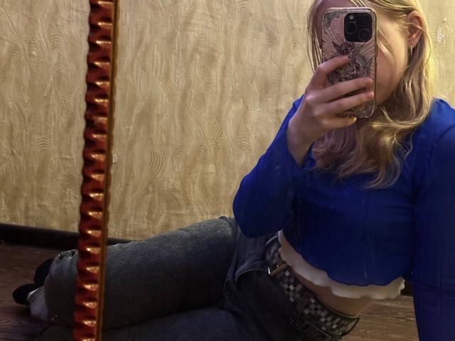 TiffaniMacandog with blonde hair, blue crop top, jeans, and belt, sitting on floor, taking mirror selfie, phone covering face, with textured wooden wall and spiral mirror stand.