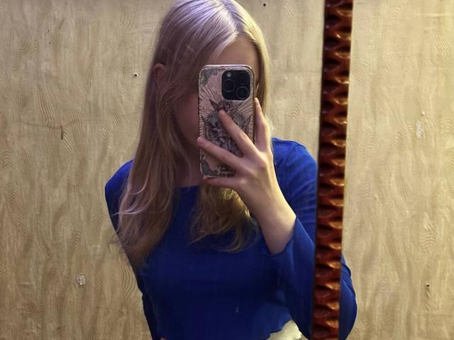 TiffaniMacandog with blonde hair, blue long-sleeve shirt, holding phone with patterned case, covering face, mirror selfie, wooden wall, visible brown textured panel on right.