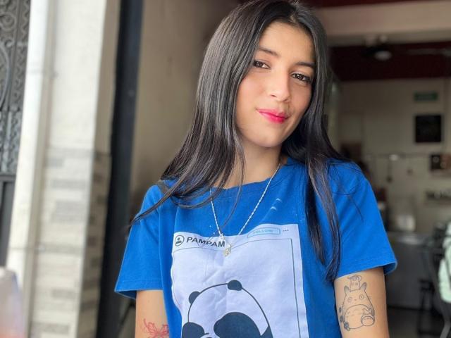 JasminKooper, con lunghi capelli neri, una camicia blu, un tatuaggio carino sul braccio destro, rossetto rosa, una collana, che sorride leggermente, in piedi di fronte a un edificio..