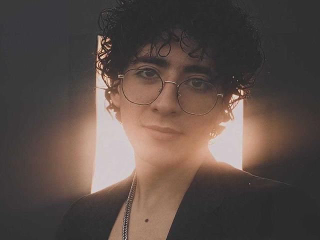 ElliotNoir with curly black hair, wearing round glasses, and a black shirt, has a small mole on their chin and a silver chain, standing in front of bright light.
