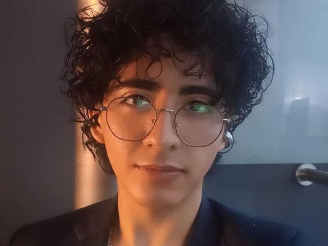 ElliotNoir with curly black hair, wearing round glasses, small hoop earrings, and a dark shirt, has a slight smile and greenish eyes in a dimly lit photo.