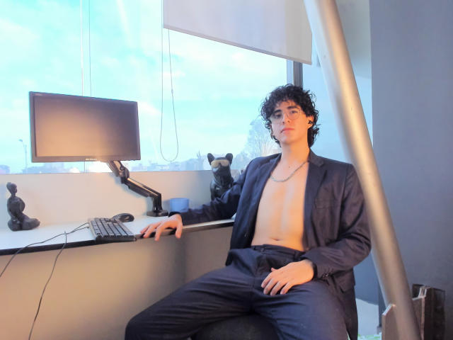 ElliotNoir with curly black hair, wearing an open blue suit, no shirt, sitting in front of a computer, hands on keyboard, looking at camera, city skyline in background.