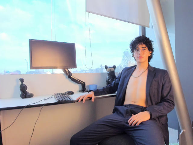 ElliotNoir with curly hair, wearing a dark blazer and no shirt, sitting by a desk with a monitor, keyboard, and mouse, looking at the camera. Two stuffed animals on desk.