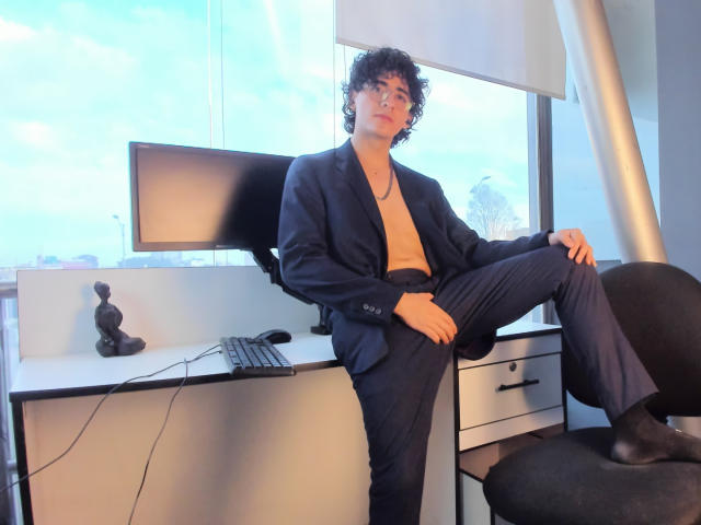 ElliotNoir with curly black hair, wearing a blue suit jacket, no shirt, and black pants, sitting with one leg up on a desk, near a computer and chair.