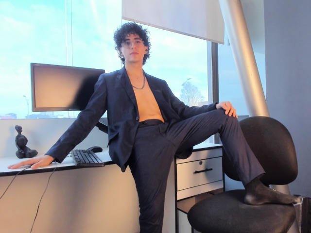 ElliotNoir with curly hair, wearing a navy suit, no shirt, black socks, and a necklace, sits with one leg on a chair, leaning on a desk, in an office.