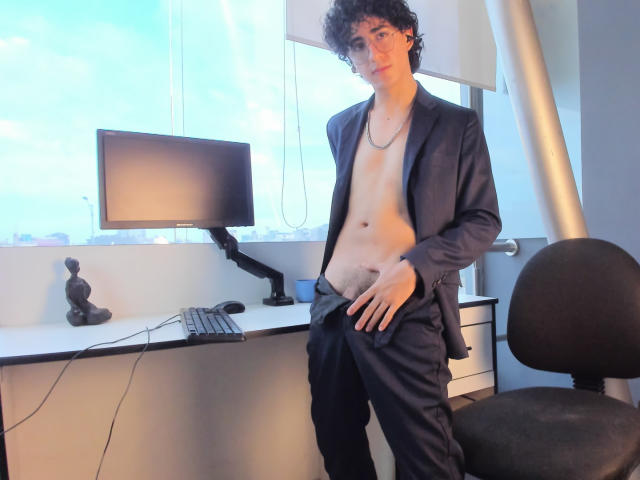 ElliotNoir, with curly brown hair, stands shirtless in a black suit, hand in pants, exposing his flaccid, uncircumcised, slightly hairy penis, in front of a desk and chair.