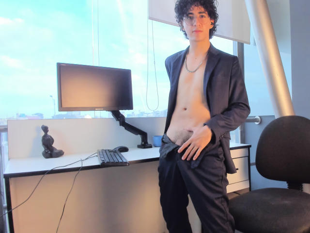ElliotNoir, with curly dark hair, stands in a room wearing an open black suit, exposing a bare, slim, flat-chested, and flaccid-ass torso, with pants slightly pulled down.