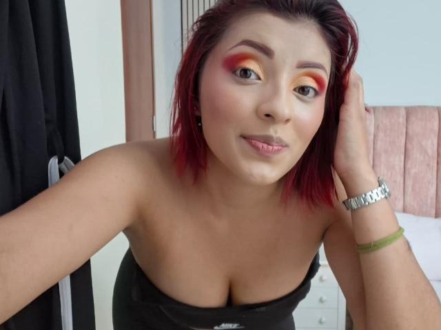 LizBonnet with red hair, orange-red eyeshadow, and pink lipstick, leaning forward, showing her tits, wearing a black strapless top, silver bracelet, and green wristband.