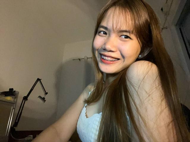 NildaReasonover with long brown hair, smiling, showing teeth, wearing a white tank top, in a dimly lit room, has a cute, attractive face and small, perky tits.