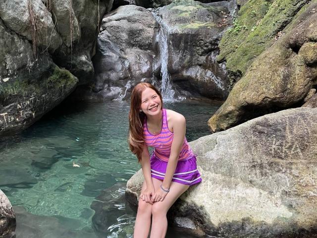 NildaReasonover with red hair, smiling, sitting on rocks, wearing a purple and pink striped dress, hands on knees, water behind her, green and brown rocky surroundings.