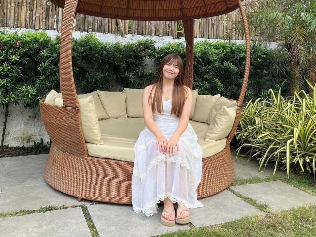 NildaReasonover sits on a wicker swing with beige cushions, wearing a white lace dress and pink sandals, smiling with long brown hair, and small tits visible under the dress.