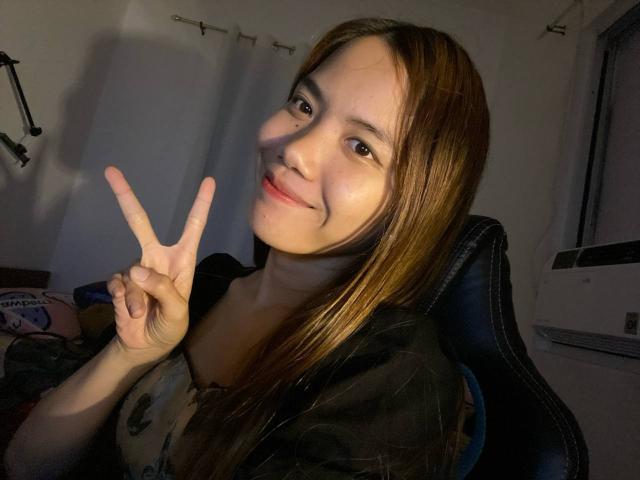 NildaReasonover with long brown hair, smiling, holding up a peace sign with her right hand, wearing a black shirt, sitting in a dark chair, indoors at night.
