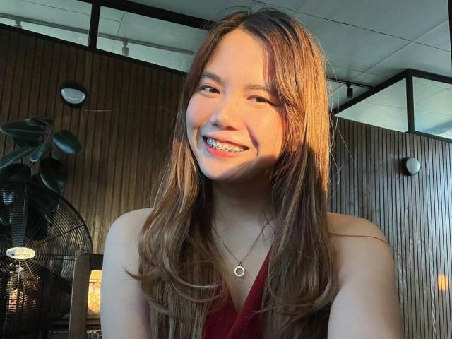NildaReasonover with long brown hair, smiling with teeth showing, wearing a red sleeveless top, and a small necklace, standing in front of a fan, in a wooden room.