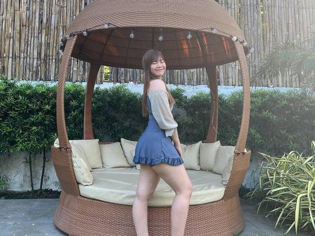NildaReasonover stands in a wicker hammock, wearing a blue off-shoulder dress, showing her ass and bare legs, with long brown hair and a smile.