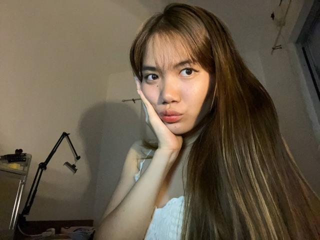 NildaReasonover with long brown hair, pouty lips, and a white tank top, resting her face on her hand, brown eyes looking at the camera, in a dimly lit room.