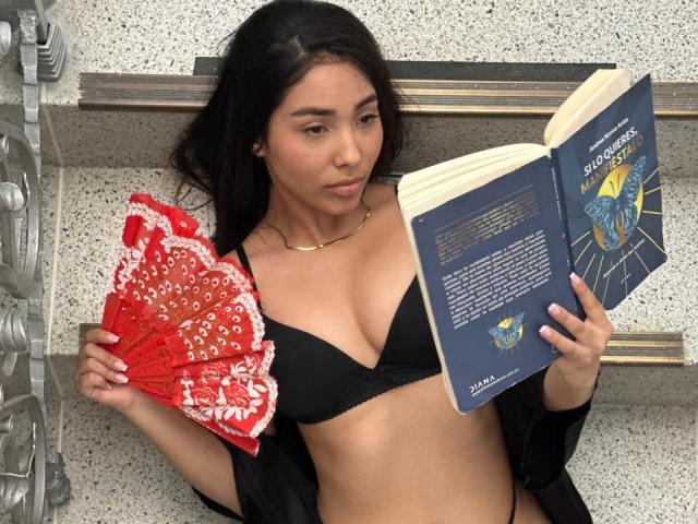SofiaCardenas with brown hair, black bra, holding red fan, and blue book, showing her tits, wearing black robe, gold necklace, in a textured, speckled, stone-like background.