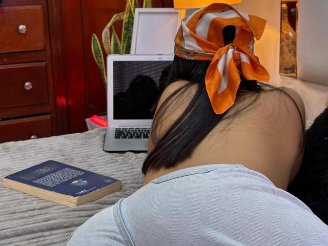 Sofia Cardenas, dos au dos de la caméra, portant une chemise grise et un foulard orange, appuyée sur un ordinateur portable, un livre sur le lit, une commode en bois en arrière-plan..