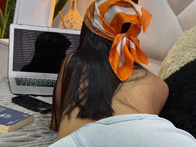 SofiaCardenas with long black hair, orange and white headscarf, lying face down on bed, laptop in front, black smartphone, book, gray and white bedding, butt up.