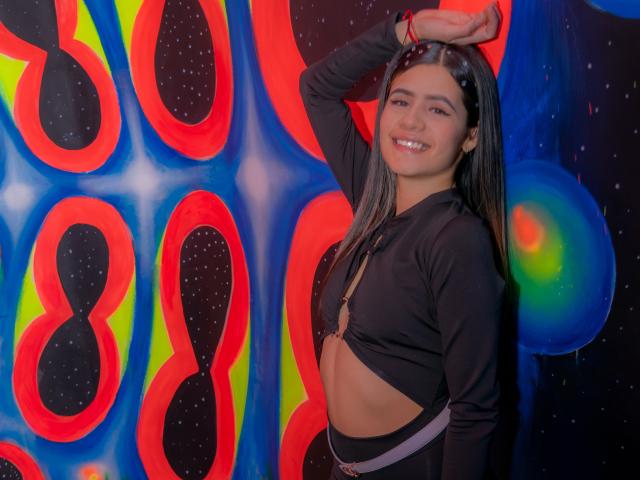 SheylaSpooner with long black hair, smiling, wearing a black long-sleeve top that exposes her small tits, standing with one arm raised, in a colorful, space-themed background.