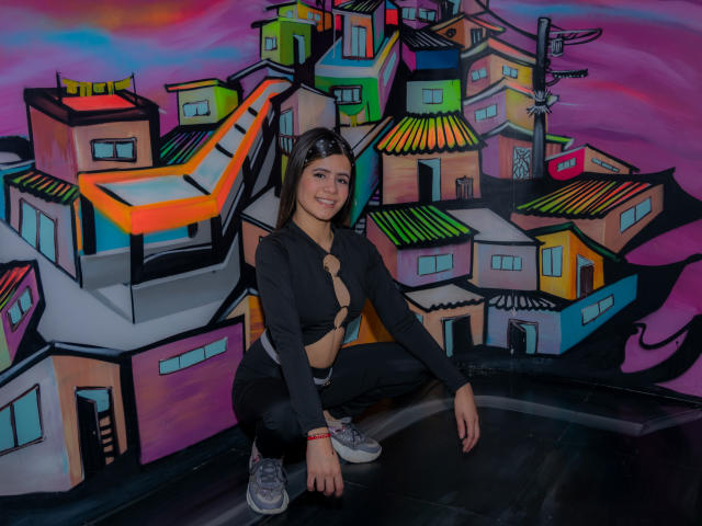 SheylaSpooner, squatting, black top with cut-out, showing small tits, black pants, gray sneakers, smiling, against colorful graffiti of urban buildings, purple and pink sky.