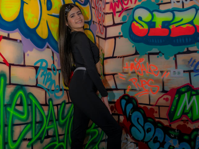 SheylaSpooner stands with long brown hair, smiling, wearing a black long-sleeve top and black pants, showing her ass, against a colorful graffiti wall.