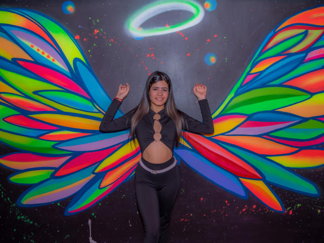 SheylaSpooner with colorful, rainbow-winged outfit, black top with cutout, black pants, smiling, arms up, neon halo above, long brown hair, black headband, standing, colorful wings.