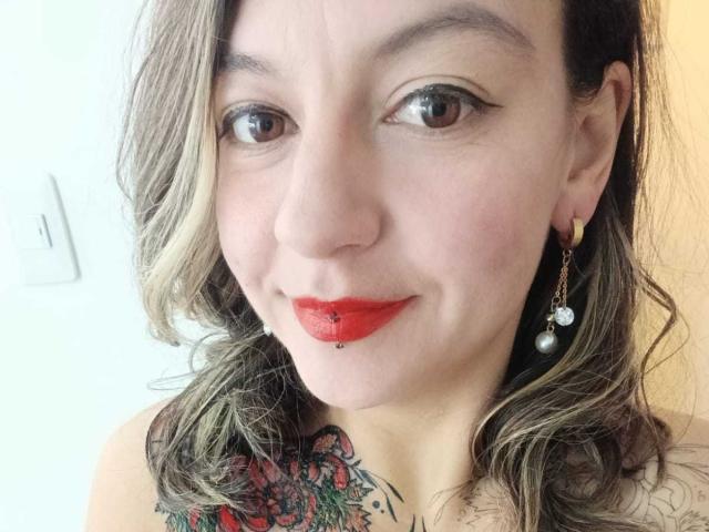 ArianaBlonde with red lipstick, nose piercing, and tattooed chest, wearing pearl earrings, smiling at the camera with wavy blonde hair, brown eyes, and fair skin.