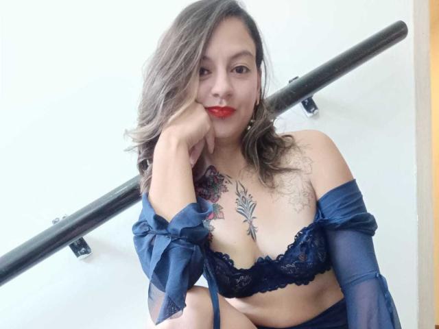 ArianaBlonde with red lipstick, blue lace bra, and sheer sleeves, leaning on a black railing, showing tattoos on chest and arm, wavy brown hair, smiling.