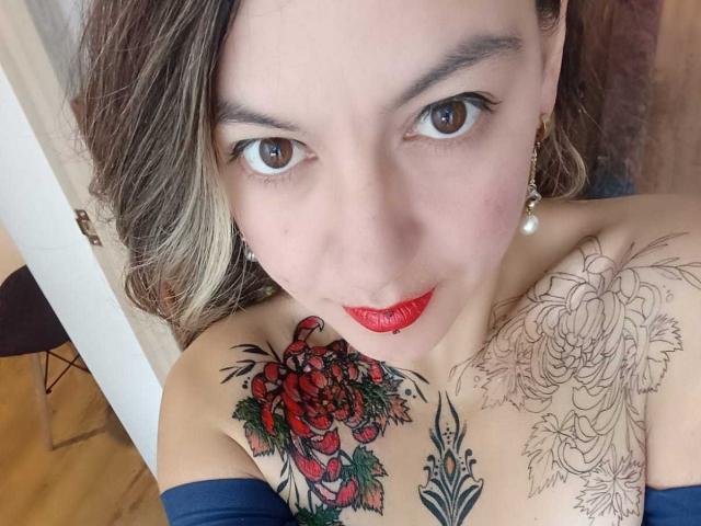 ArianaBlonde with tattoos, red lipstick, and blue sleeves, looking up with brown eyes, pearl earrings, and blonde hair, showing a rose and flower tattoo on her tits.