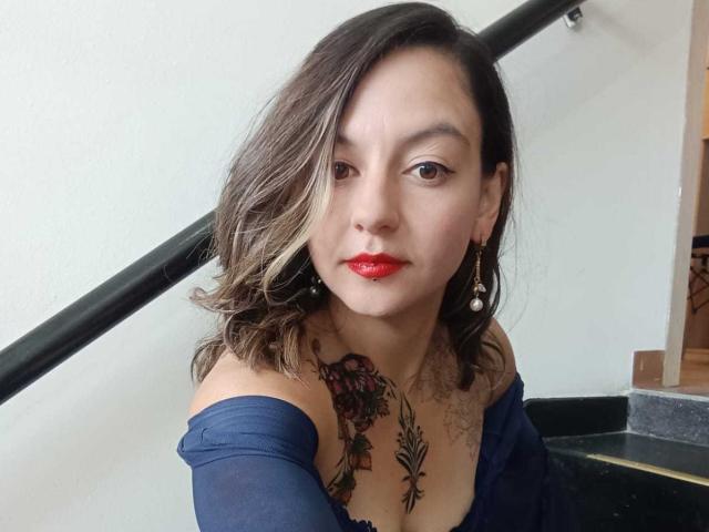 ArianaBlonde with shoulder-length brown hair, red lipstick, and blue off-shoulder top, showing tattoos on her chest and neck, standing by a black staircase.