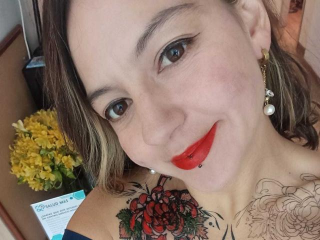 ArianaBlonde has red lipstick, a lip piercing, and a tattoo of red roses on her shoulder. She's smiling with brown eyes and wearing pearl earrings.