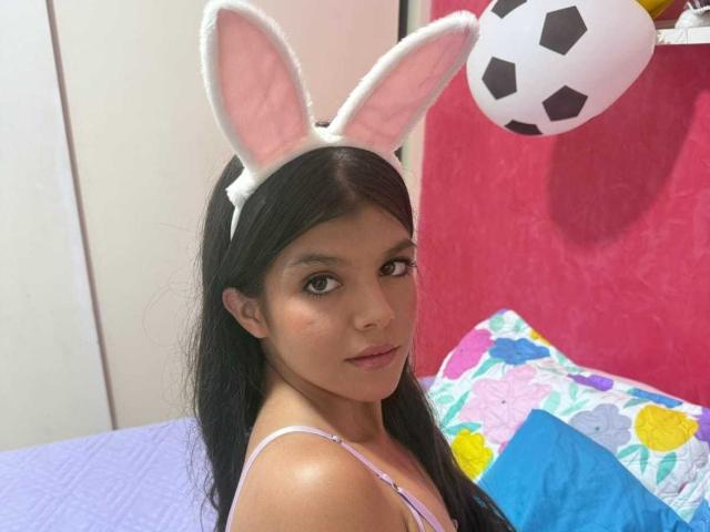 RilleyFoyx with long black hair, wearing white bunny ears, pink and white top, looking at camera, sitting on bed with colorful pillow, soccer ball hanging above.