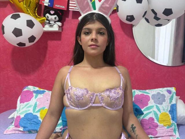 RilleyFoyx with purple lace bra, small tits, brown hair, white bunny ears, pink wall, soccer balls, colorful pillow, tattoo on right forearm, sitting on bed.