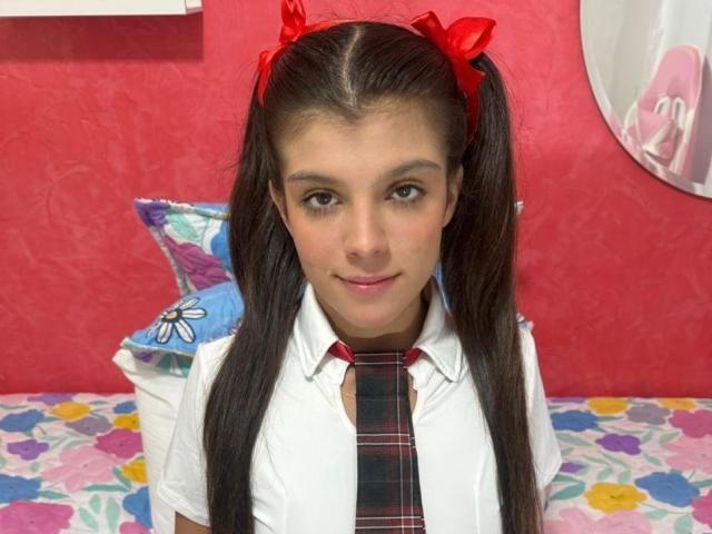 RilleyFoyx with long brown pigtails, red hair bows, white shirt, plaid tie, smiling, pink wall, floral bedspread, and blue pillow with white flower.