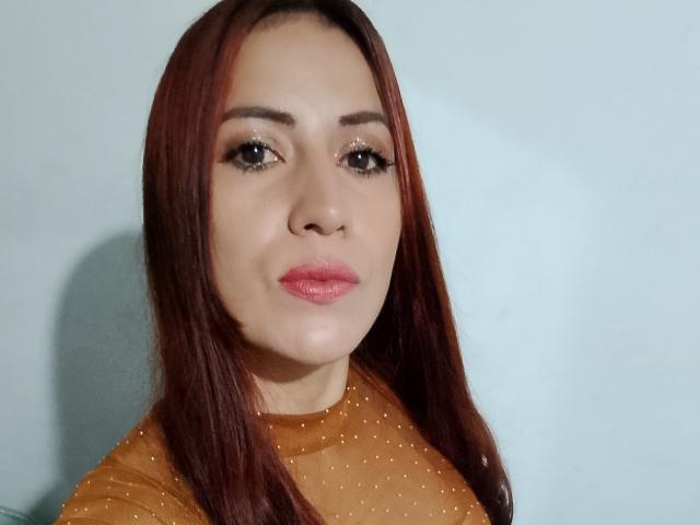 SofiaAnal, met rood haar, draagt een doorschijnend bruin topje met stippen, heeft roze lippen, subtiele make-up en een serieuze uitdrukking. Haar borsten zijn niet zichtbaar..