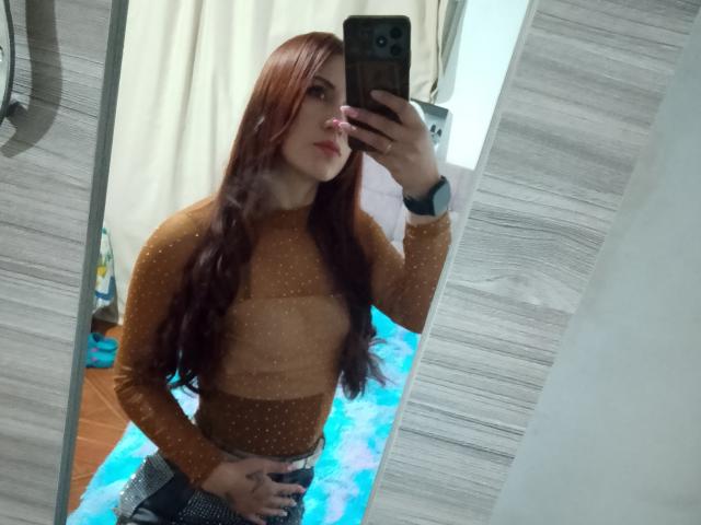 SofiaAnal with long red hair, wearing a brown, sheer, polka-dot top, taking a mirror selfie, showing her hand on her butt, in a room with a blue rug.