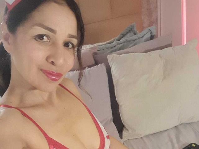 Olivia Scott, con cabello oscuro, vestida con un top de bikini rojo, labios rosados y una sonrisa encantadora, mostrando sus pequeños senos, acostada en una cama con almohadas..
