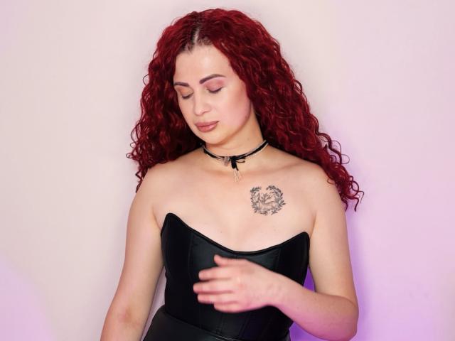 LucyGrayyy with curly red hair, wearing a black choker and strapless black top, showing a tattoo on her right boob, looking down with a slight smile.