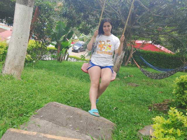 SofiaAnal with tan skin and dark hair, wearing a white "La Liga" shirt, blue shorts, and teal sandals, swinging on a rope, standing on a rock, smiling.