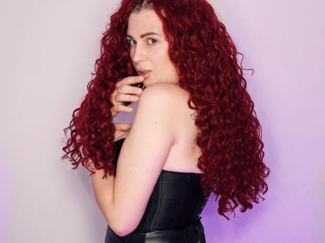 LucyGrayyy with long red curly hair, black strapless leather top, biting her finger, looking over her shoulder, pale skin, subtle tattoo on her right shoulder blade.