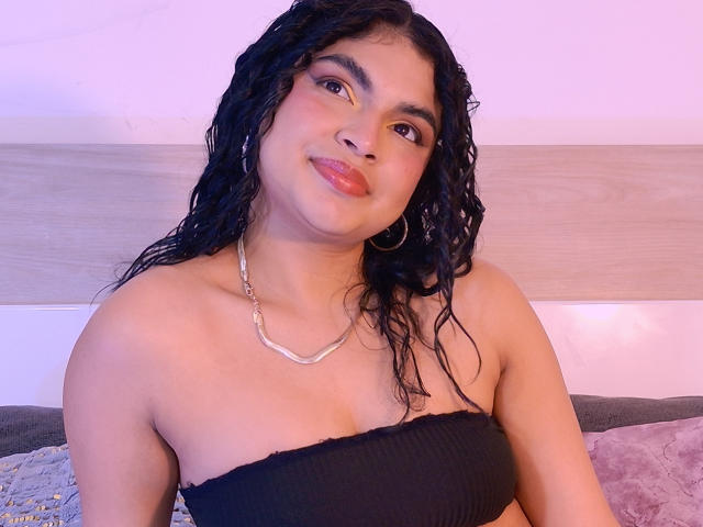 MelodyAlvarez with curly black hair, wearing a black strapless top, silver necklace, and hoop earrings, smiling with pink lipstick. Her tits are small and her skin is tan.